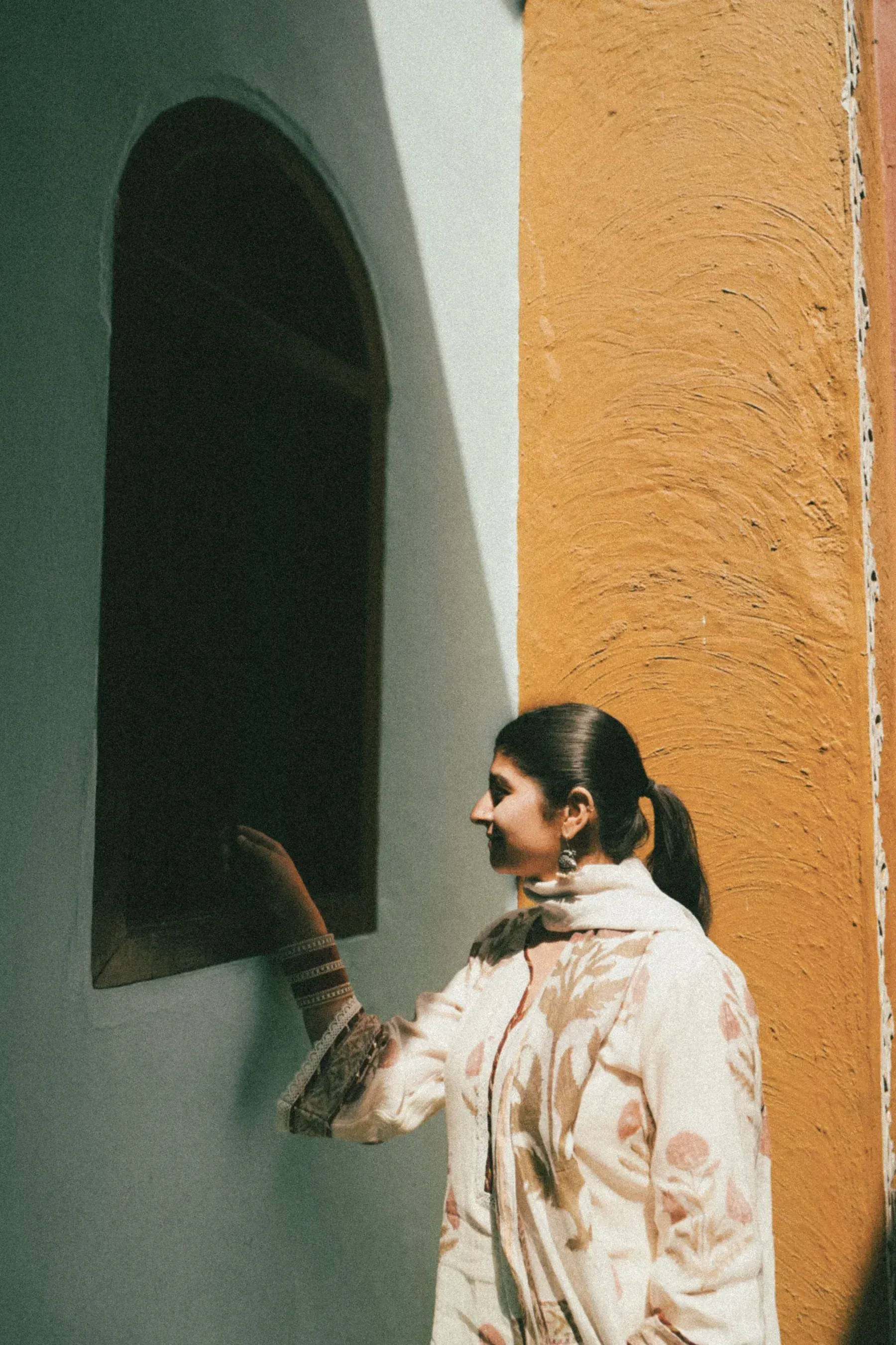 A woman in a printed ivory kurta beside a sage wall and an arched window