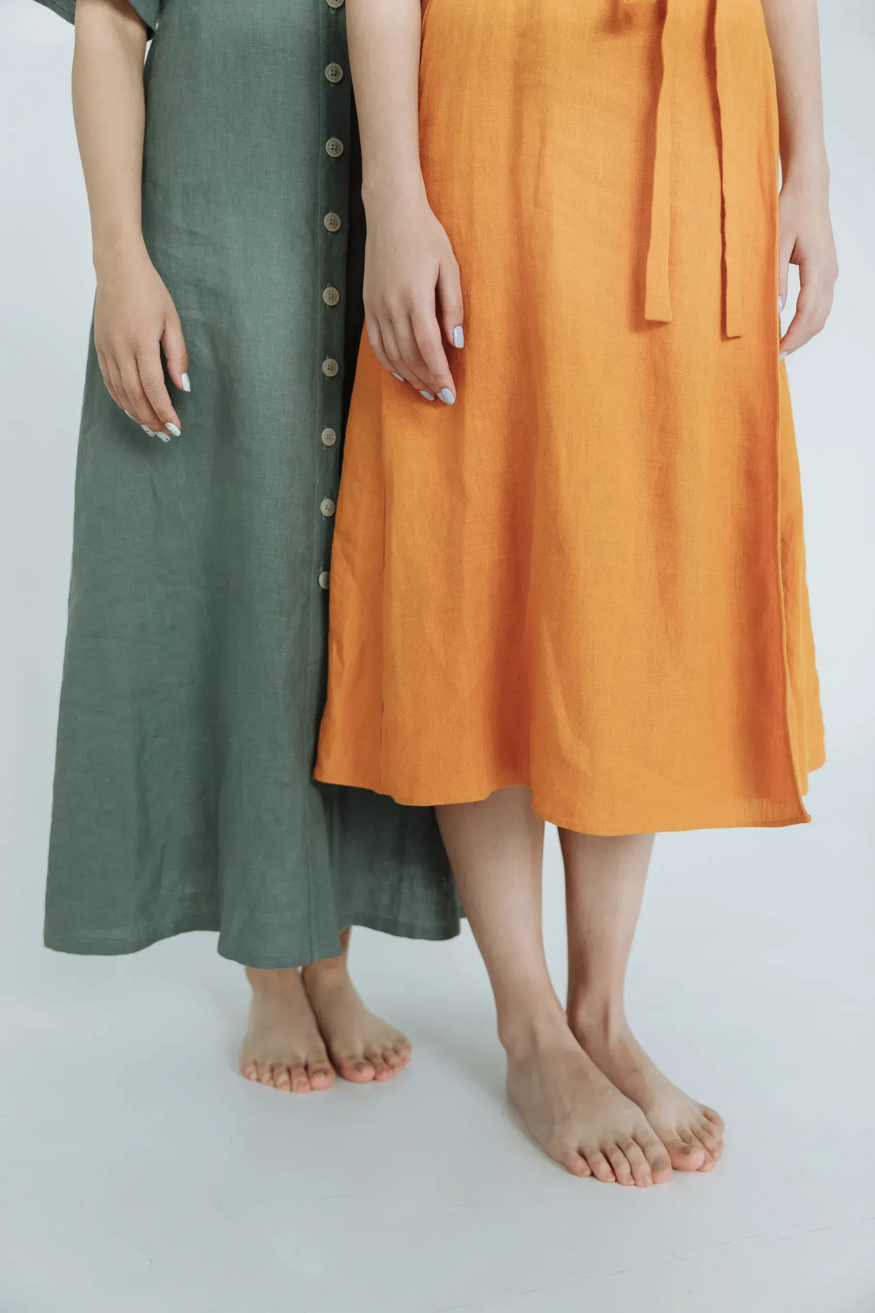 Two women in sage and marigold linen dresses, shown from the waist down