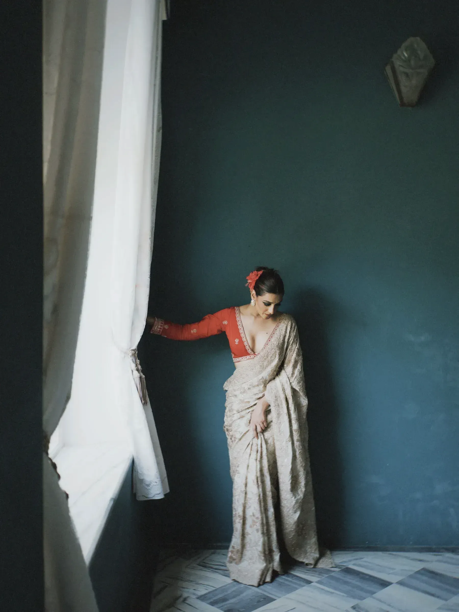 A woman in an ivory printed saree against a teal wall beside a white curtain