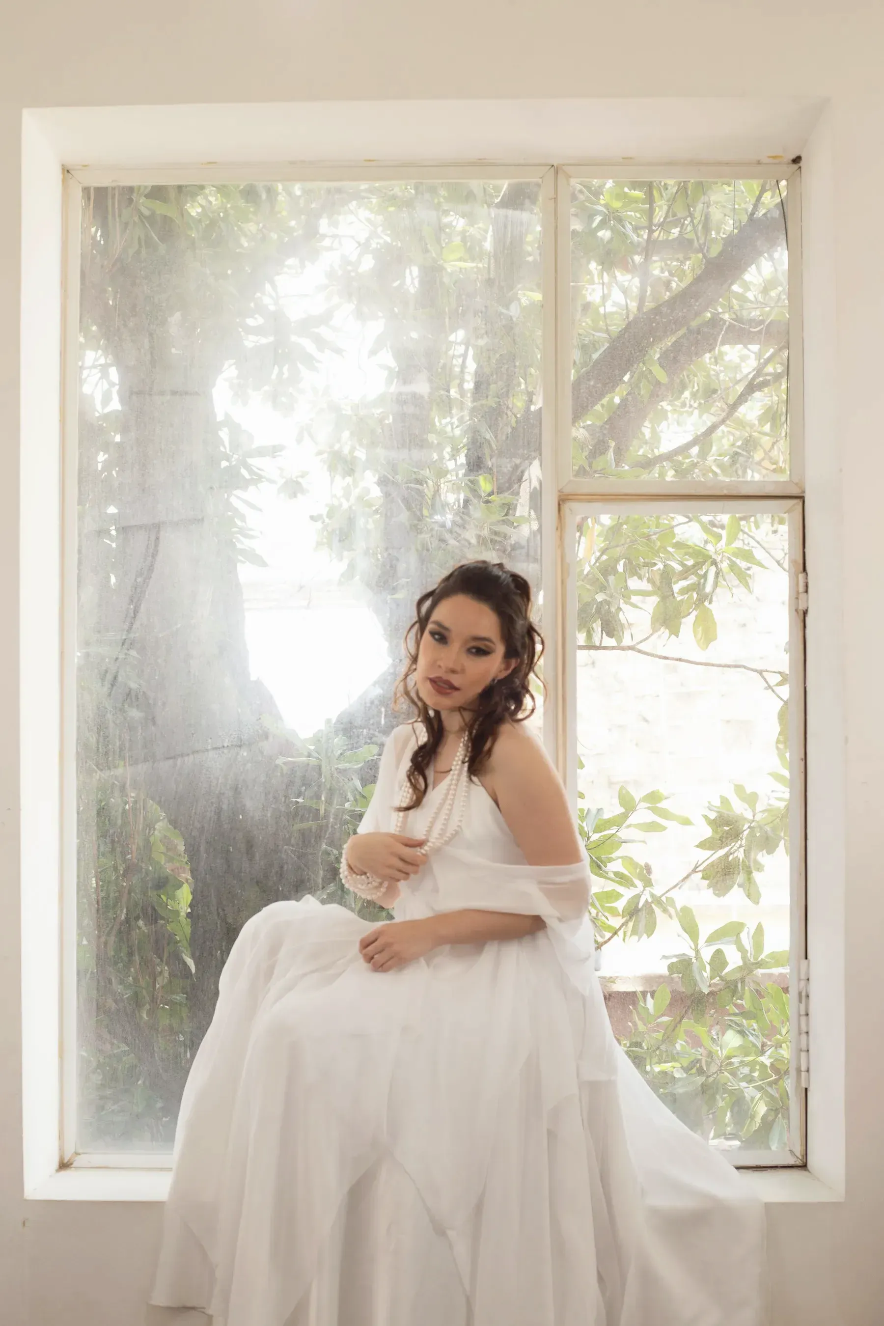 A woman in an ivory mulmul dress seated by a tall window