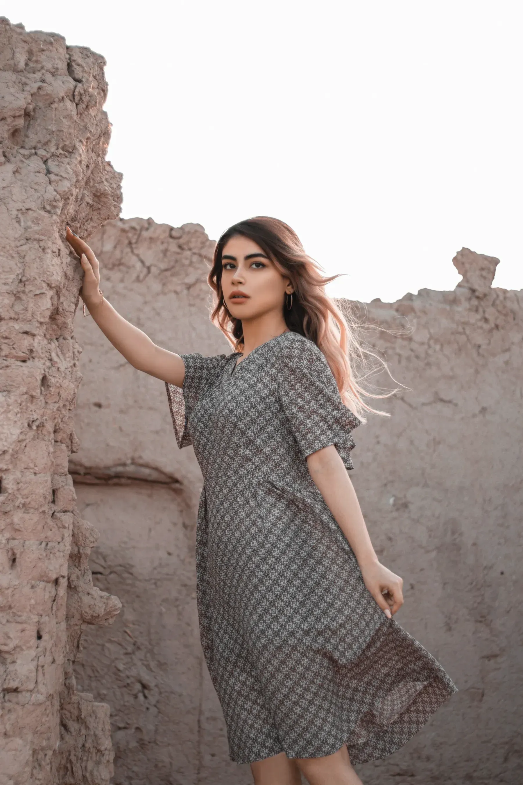 A woman in the slate midi dress leaning on a mud wall